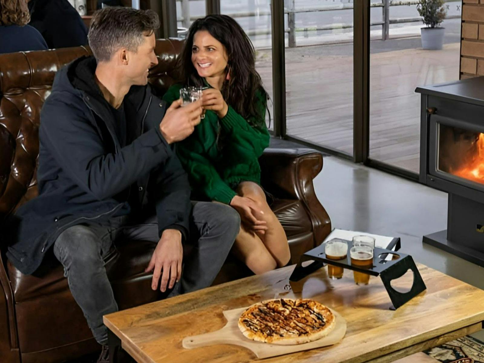 Couple drinking beer tasting paddle with pizza by lit woodfire