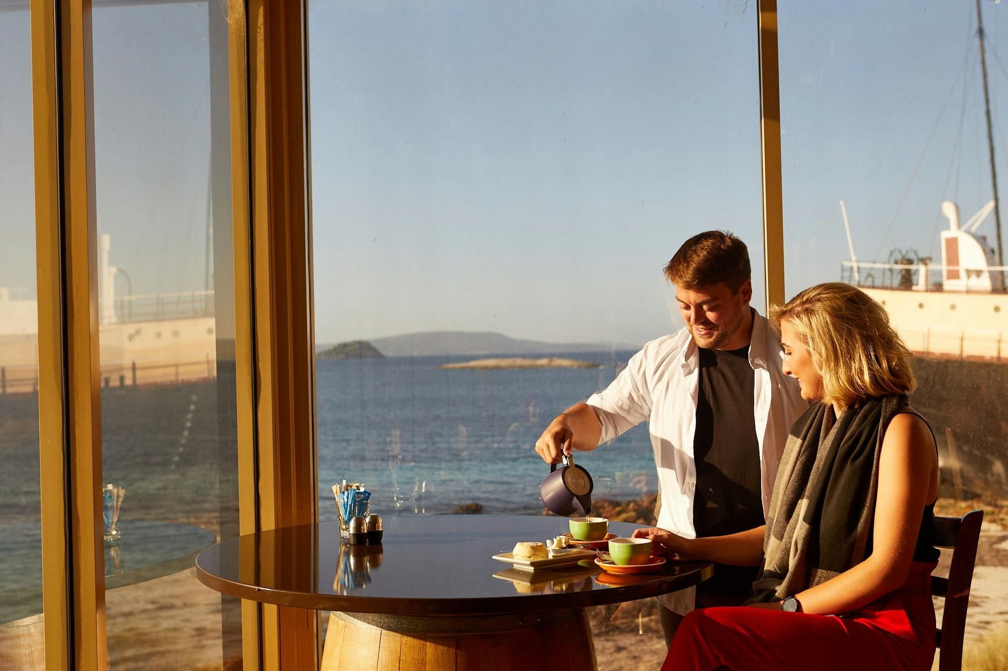 man pouring from pot into cup on table with woman lookin gon and ocean in background