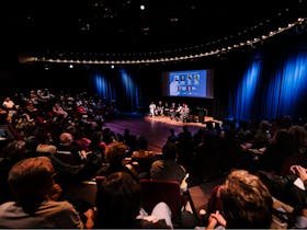 A diverse crowd is seated, listening intently to a panel discussion taking place on stage.