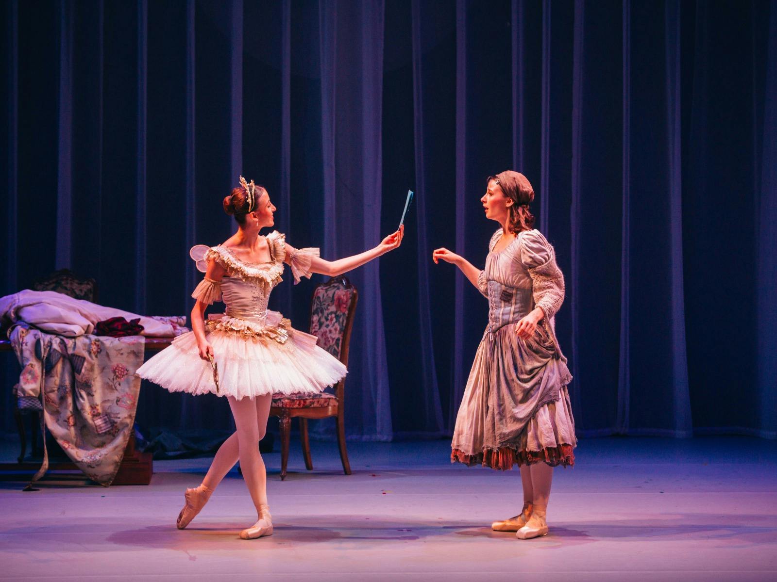 A ballet dancer in a white tutu and tiara hands a small blue object to a dancer in a peasant dress.