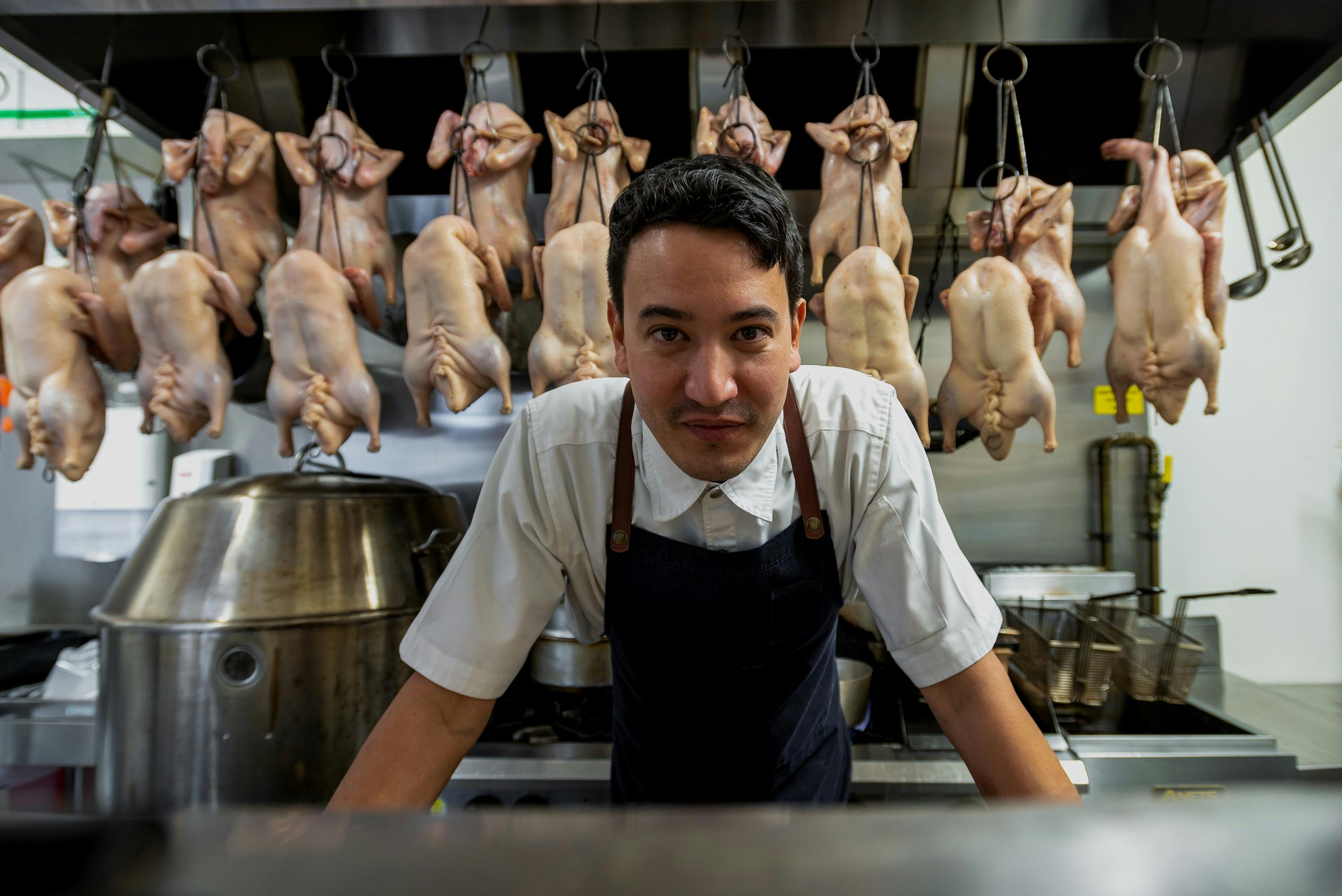 Man in Chef uniform in leaning on bench in front of lined up brined ducks hanging in kitchen