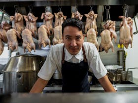 Man in Chef uniform in leaning on bench in front of lined up brined ducks hanging in kitchen