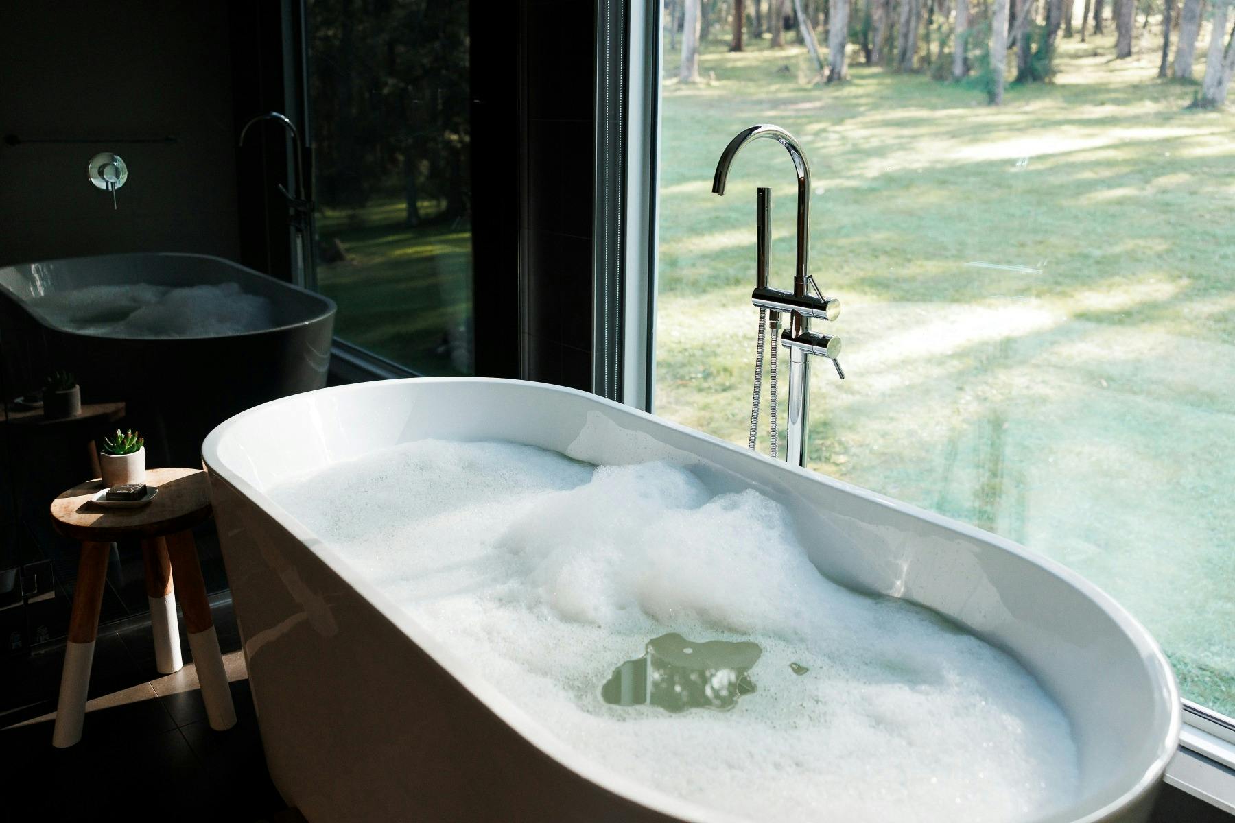 Large deep freestanding bath filled with bubbles and a large window with views of bushland