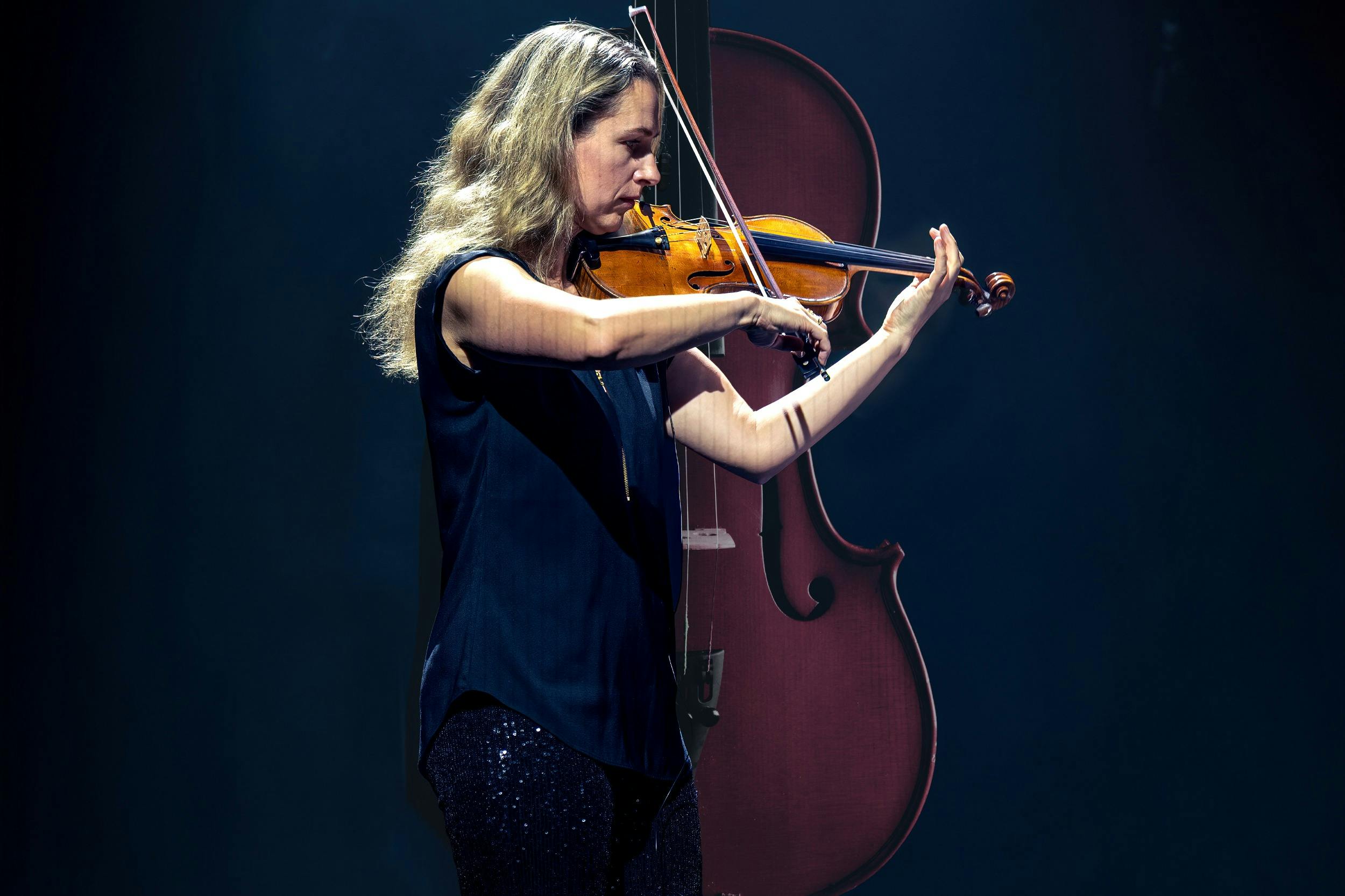Violinist Emma McGrath performing on stage under a spotlight. She has long, wavy hair