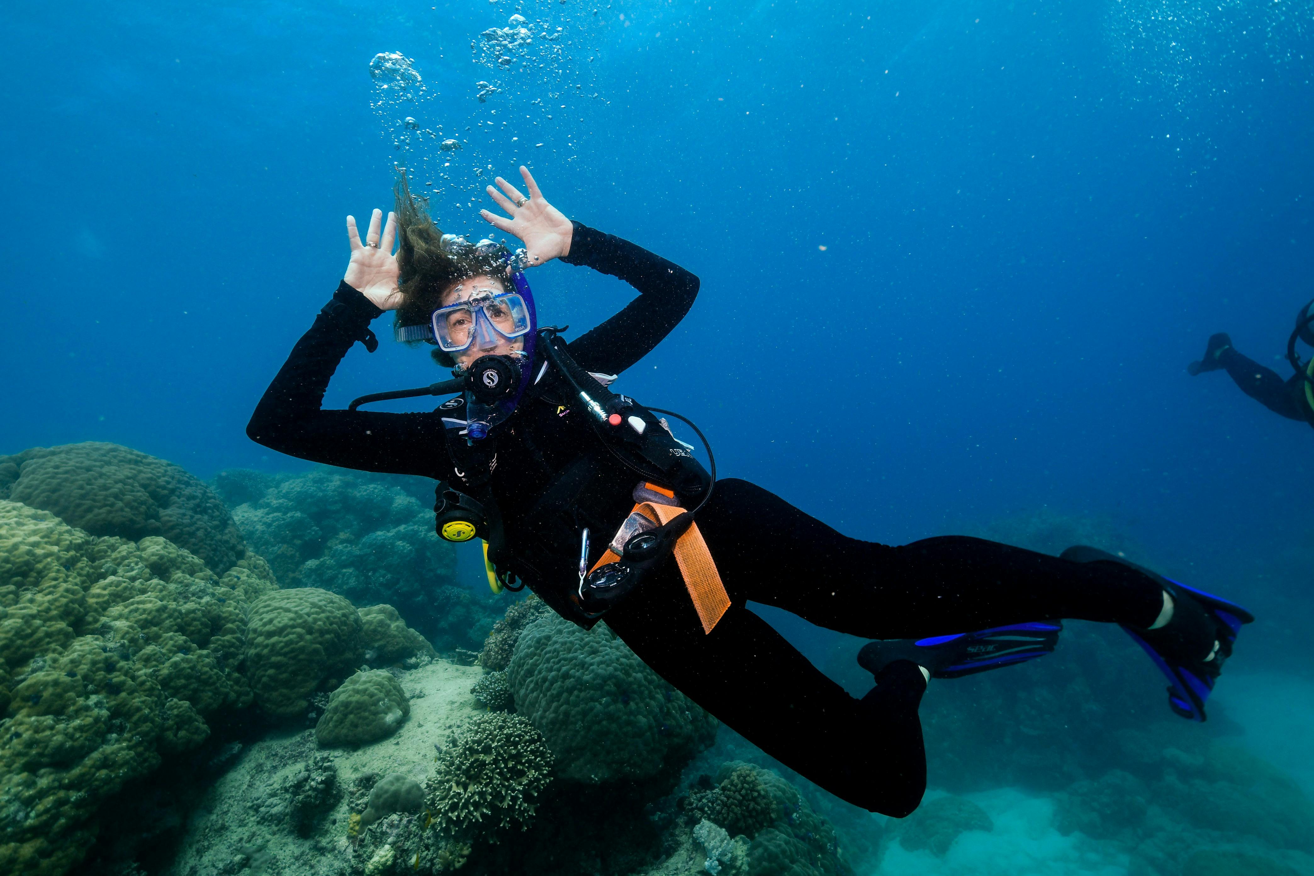 Scuba diver in blue water waving with both hands at camera with reef in the background