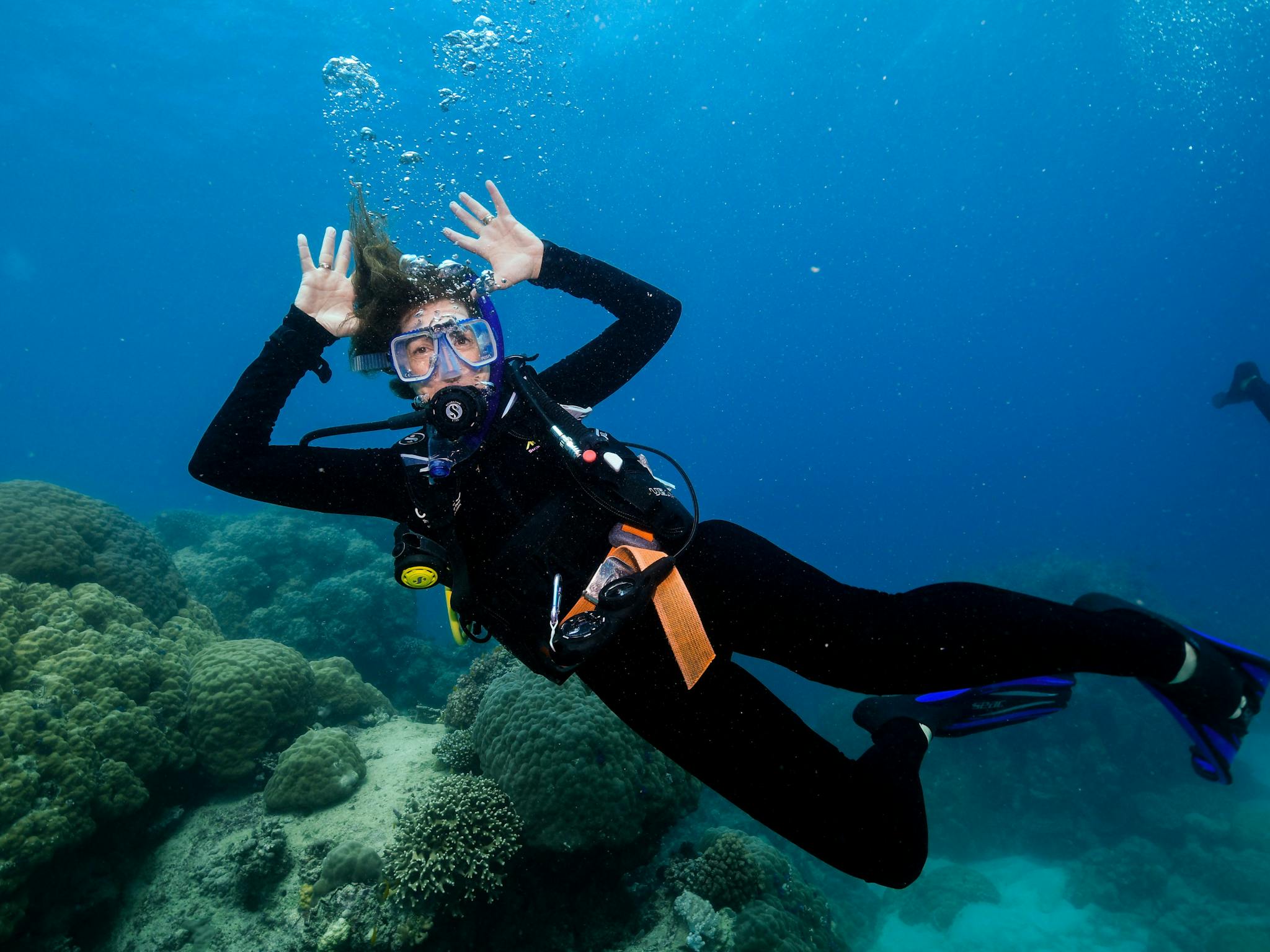 Scuba diver in blue water waving with both hands at camera with reef in the background Scuba diver in blue water waving with both hands at camera with reef in the background