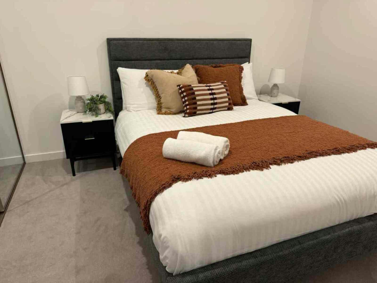 Bedroom with grey fabric headboard, white linens, rust-orange throw blanket, and black bedside table