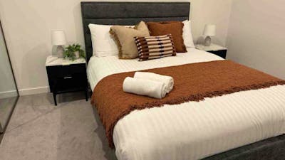 Bedroom with grey fabric headboard, white linens, rust-orange throw blanket, and black bedside table