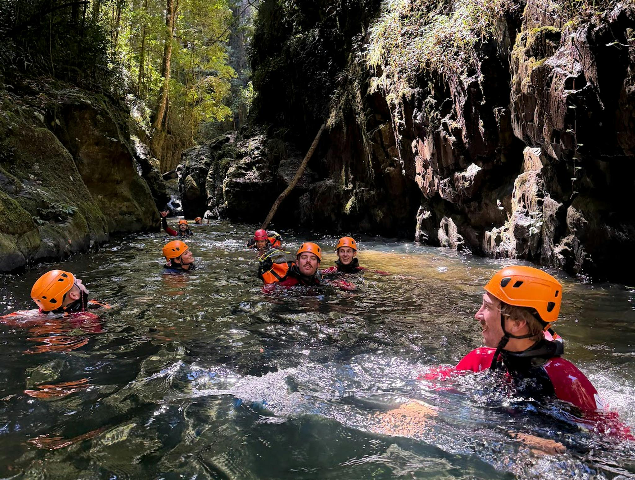 Group of canyoners in orange helmets swimming through a narrow rocky gorge in Rosewood Canyon, Dorri