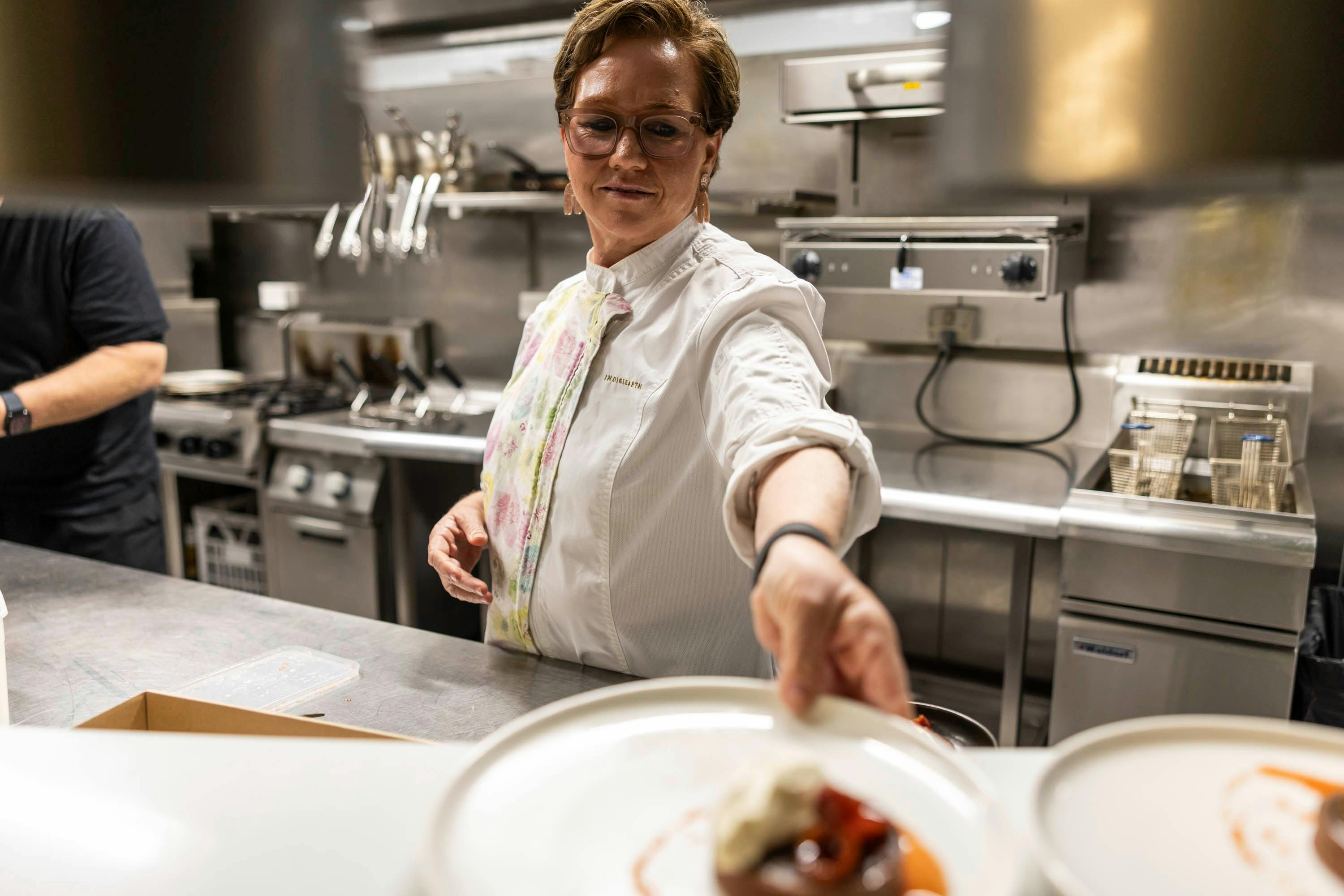 Sharon Winsor in a commercial kitchen passing a plate to a server off camera
