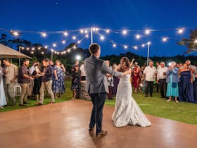 Bride & Groom enjoying their first dance, outside at dusk, under festoon lights with guests watching