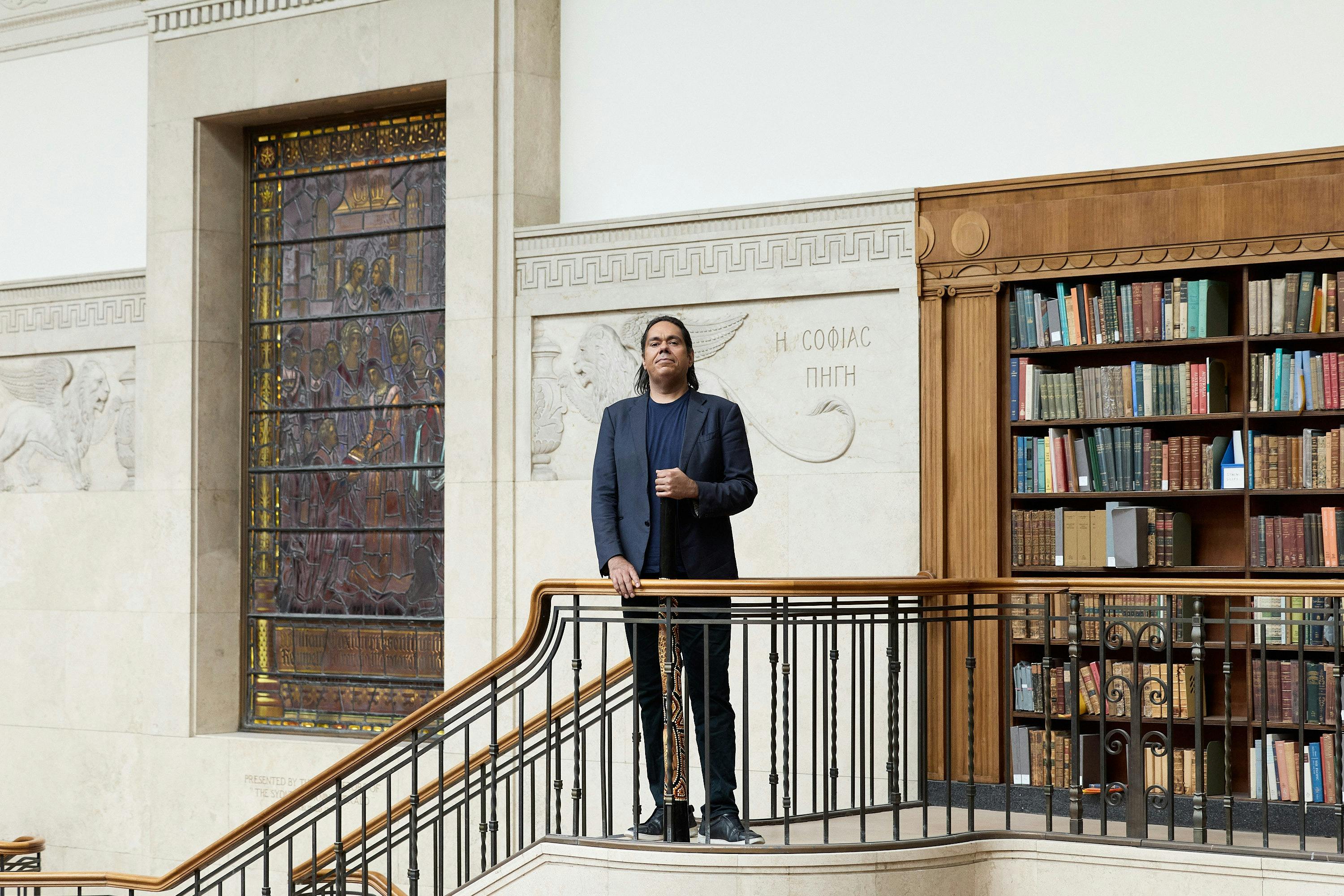 A man standing with a didgeridoo by the staircase in a library