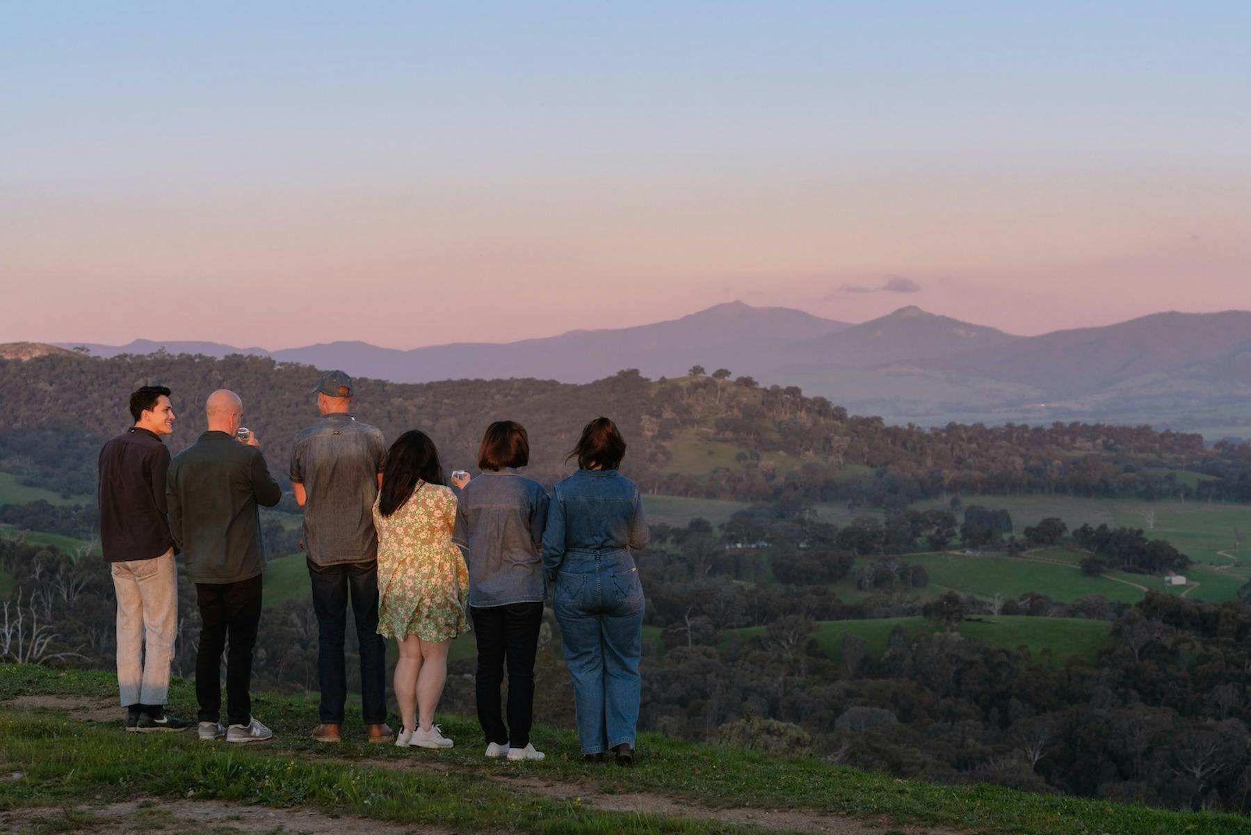 A group of people stand together on a hilltop, holding drinks and watching the sunset.