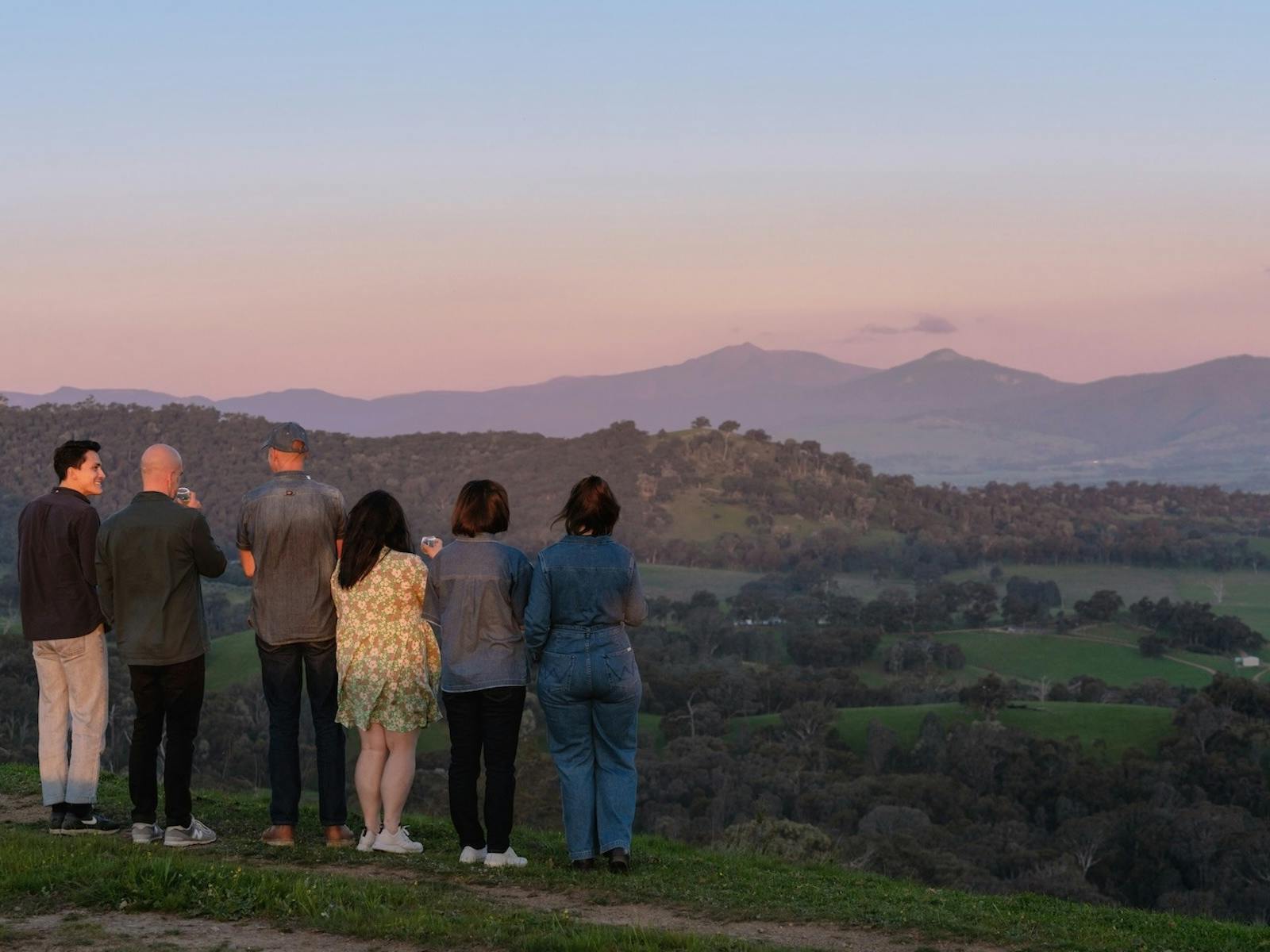 A group of people stand together on a hilltop, holding drinks and watching the sunset.