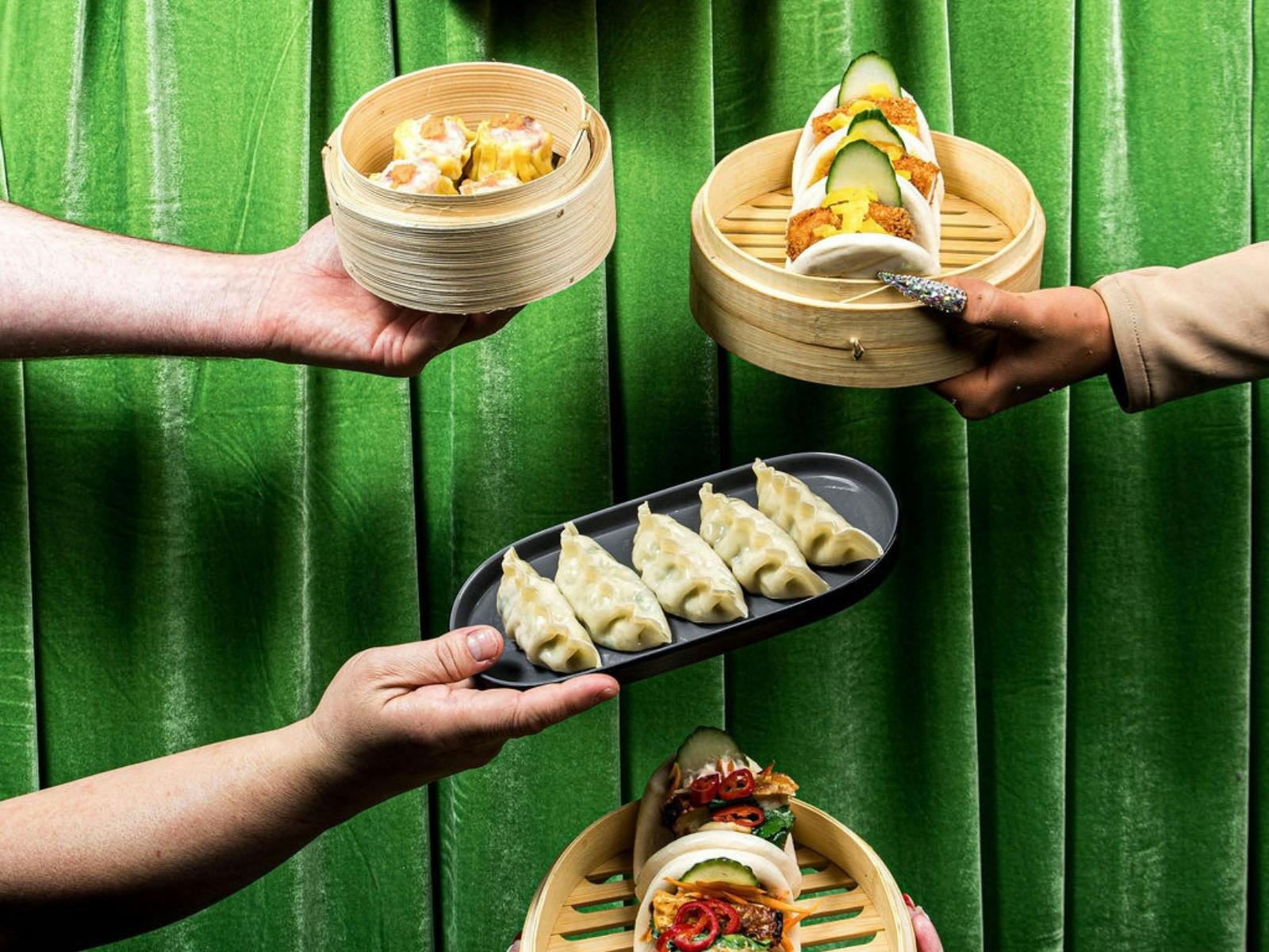 Hands hold bamboo steamers of bao and dumplings against a green curtain.