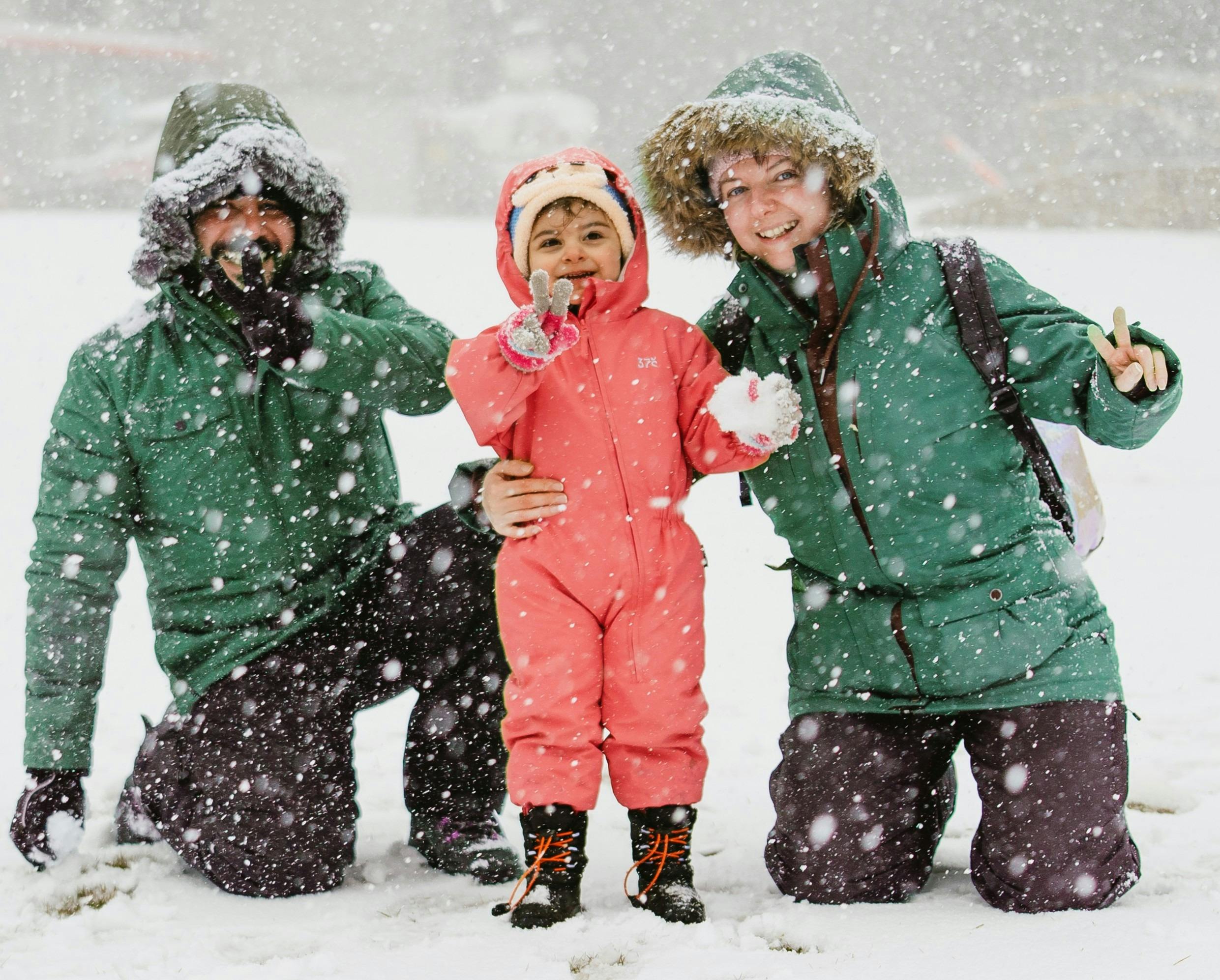 Two adults kneeling in the snow next to a young child while it is snowing