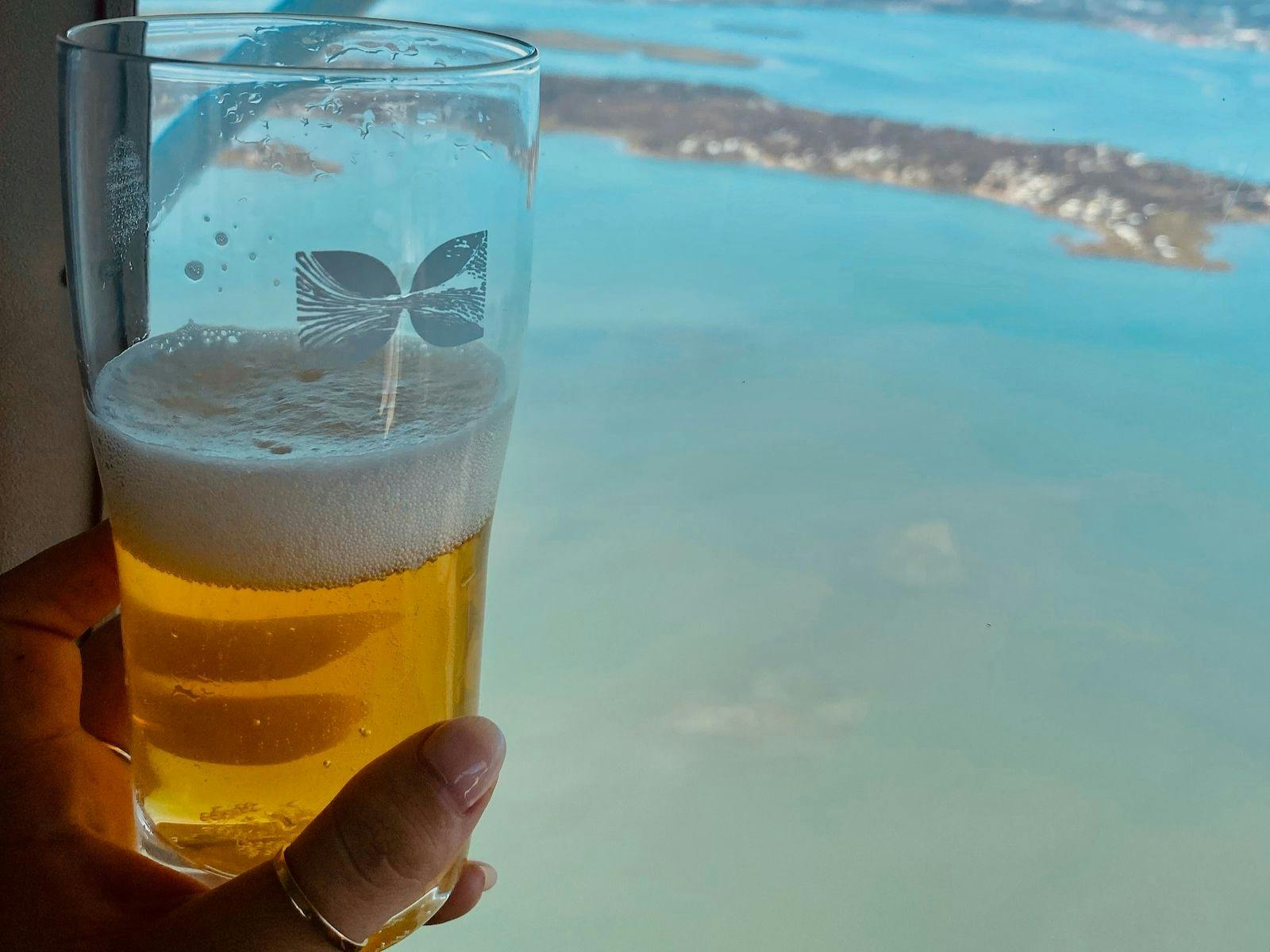 A glass of beer with logo in the foreground, held by a hand. Background is blue water and landmass