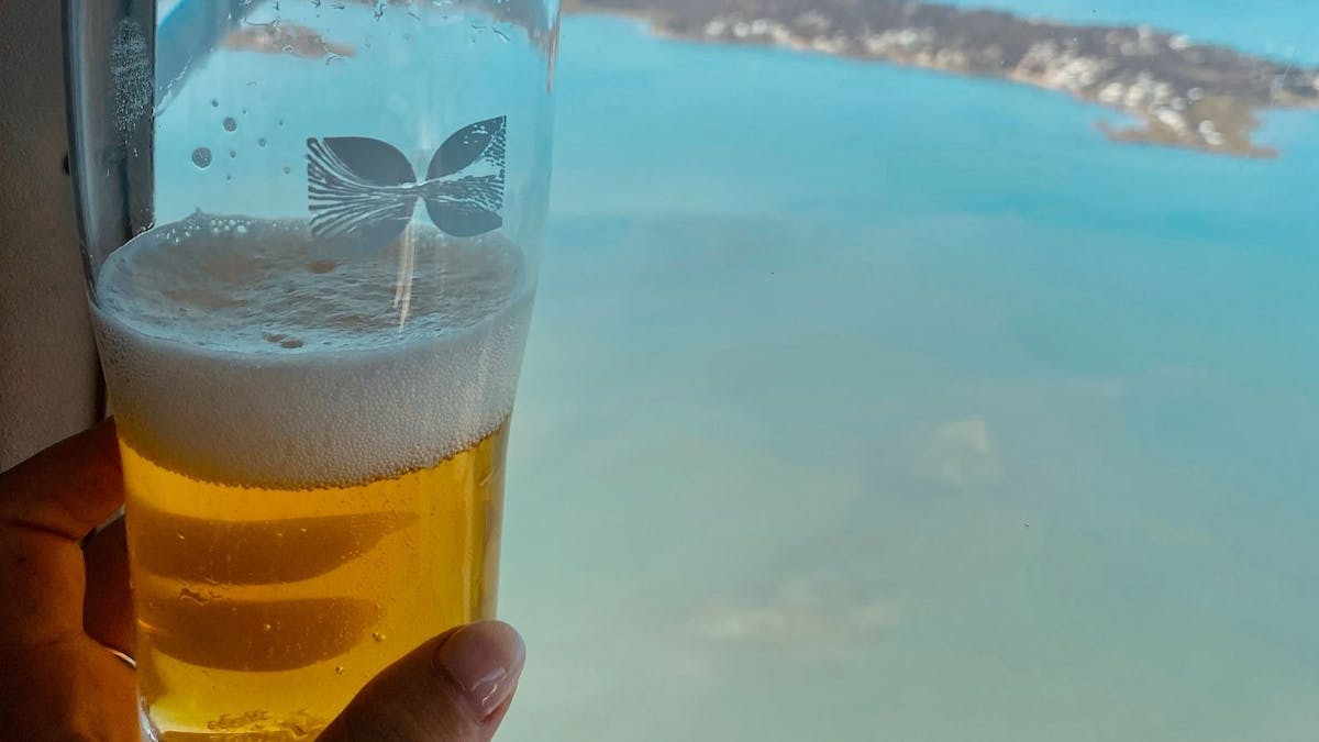A glass of beer with logo in the foreground, held by a hand. Background is blue water and landmass