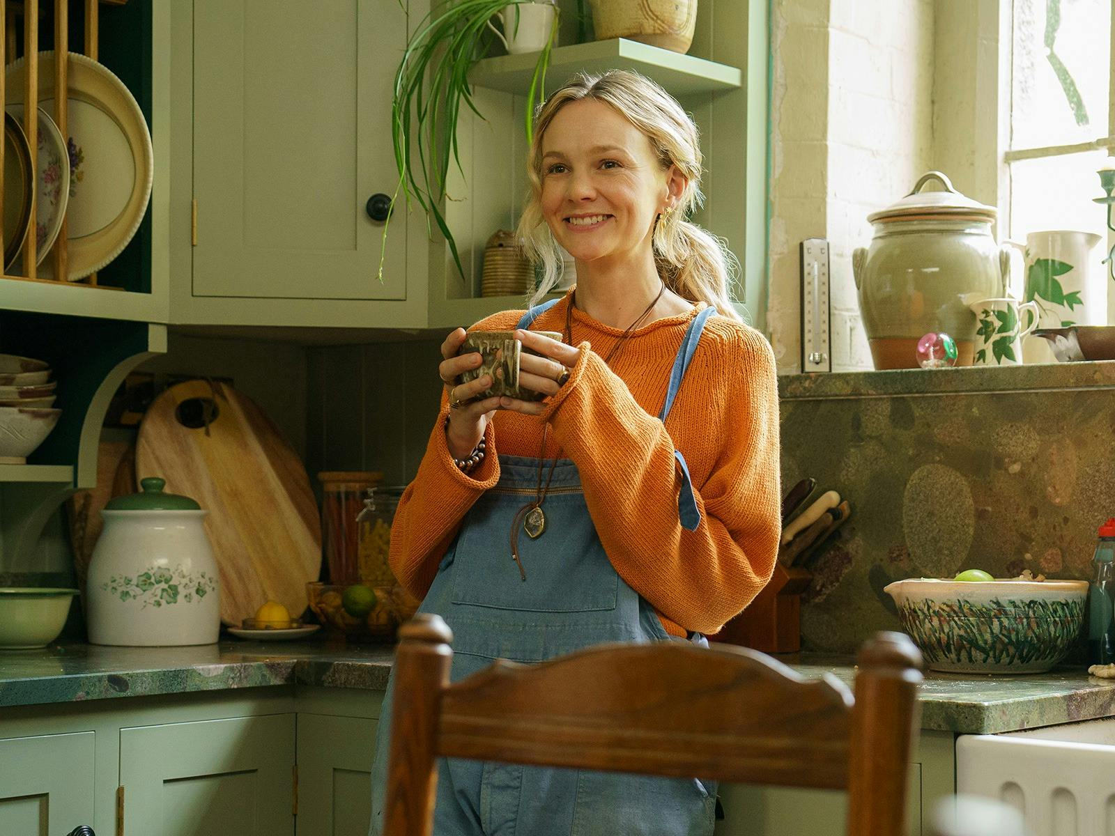 A young woman stands in the kitchen holding a mug, smiling.