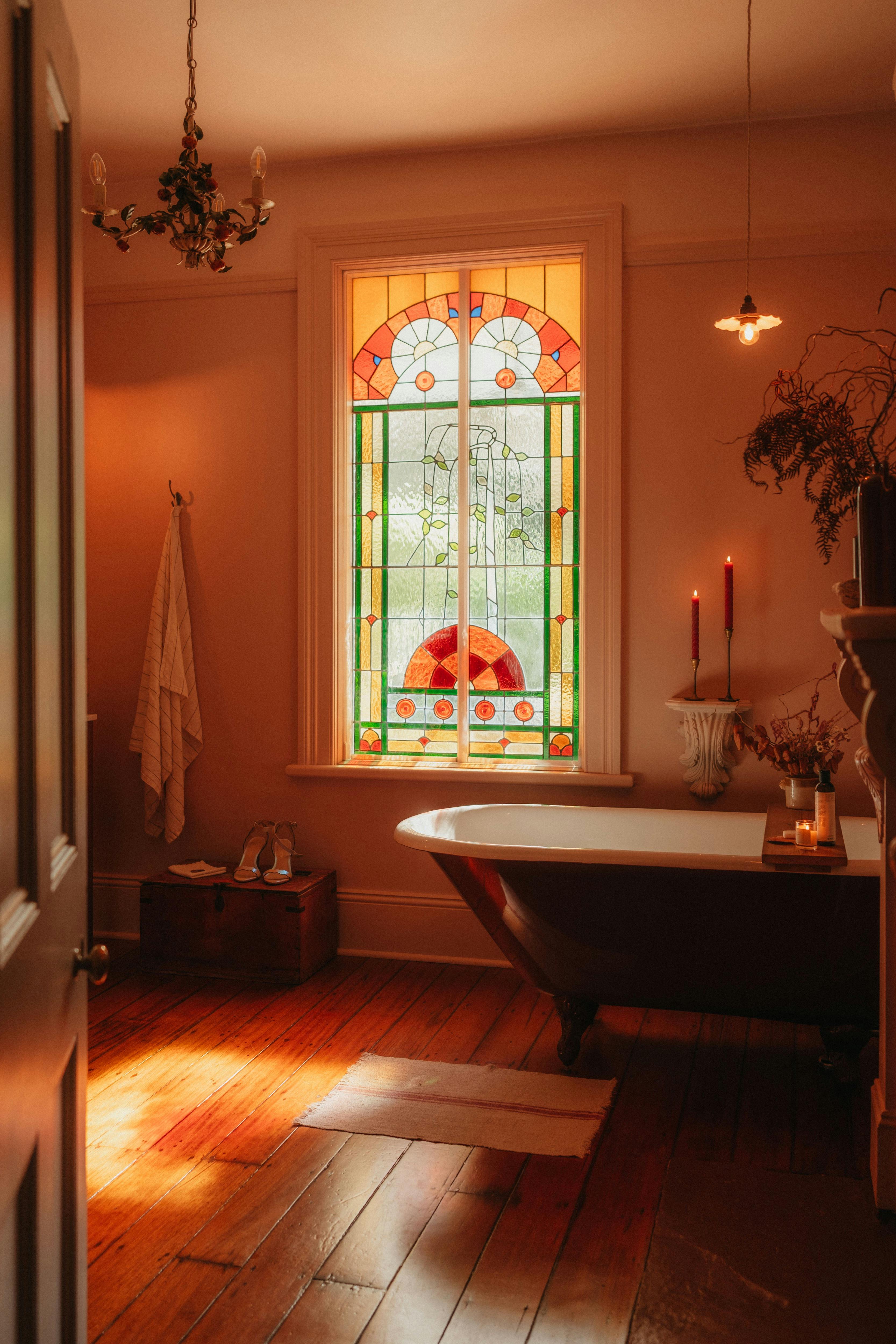 Clawfoot bath beneath stained glass window in warm, softly lit bathroom.
