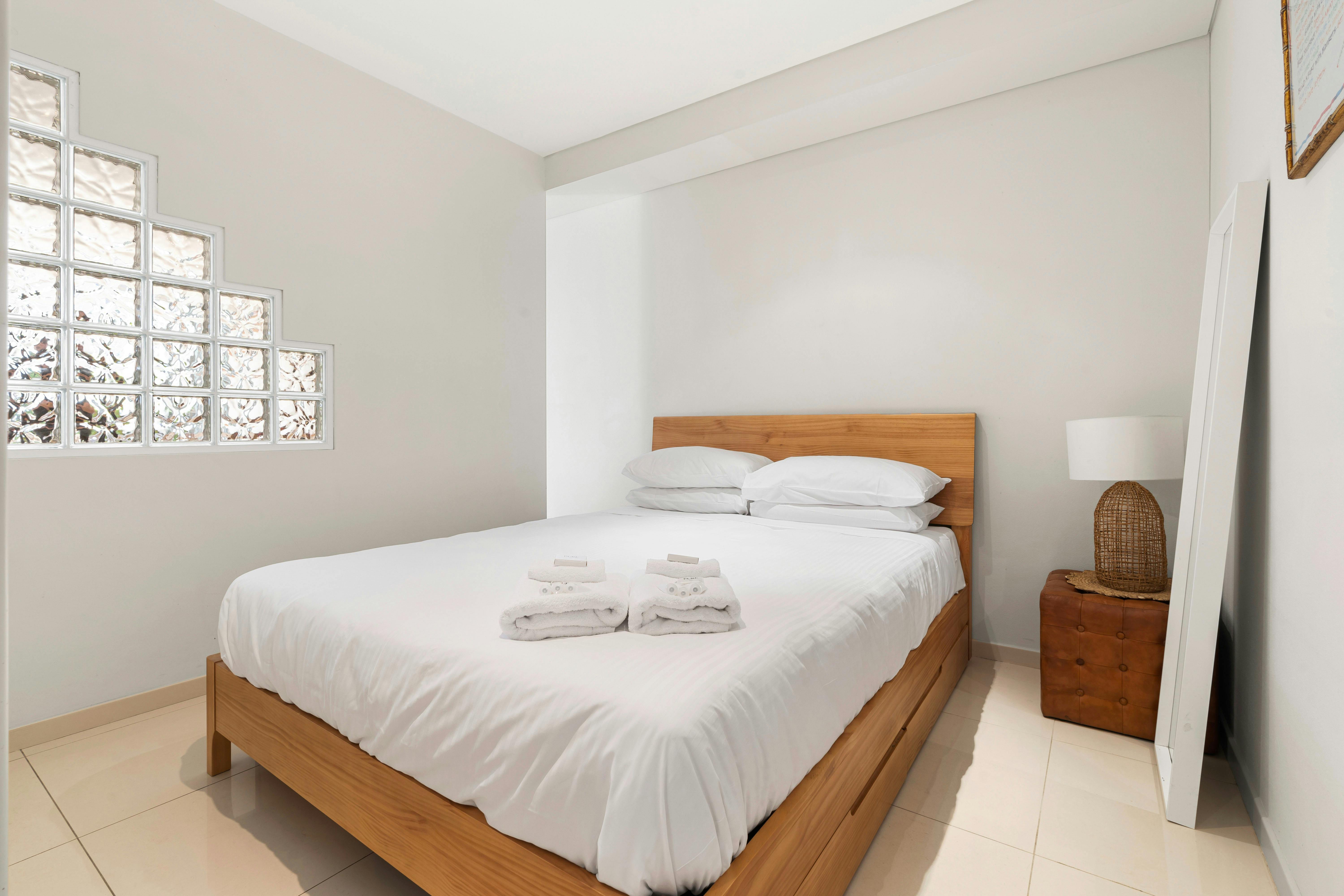 Bright bedroom with a wooden bed, soft linens, and unique glass block window feature.