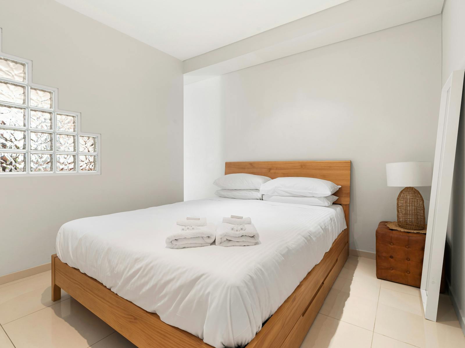 Bright bedroom with a wooden bed, soft linens, and unique glass block window feature.