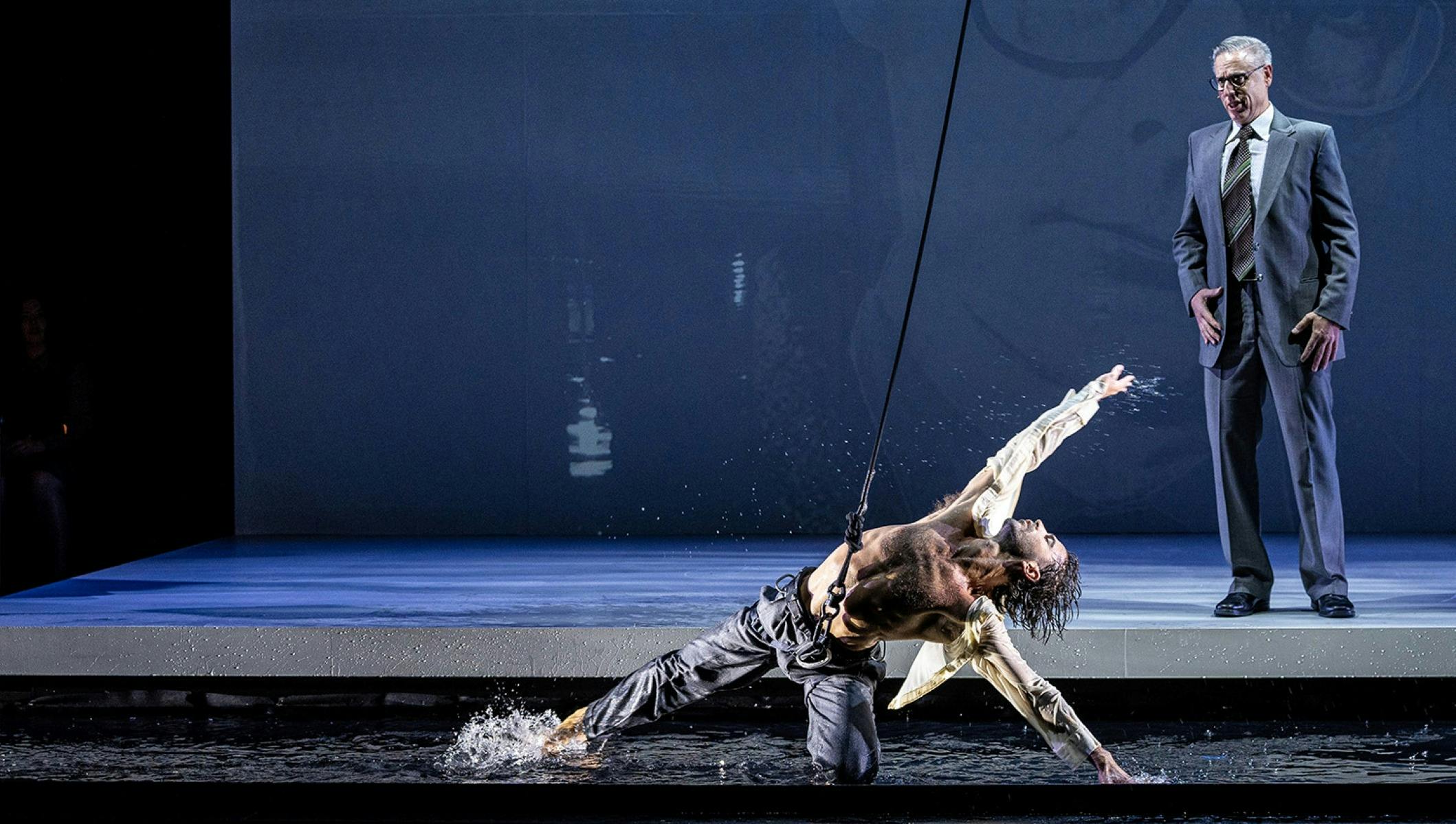 Performer suspended over water reaches out dramatically as another stands on a bare stage.
