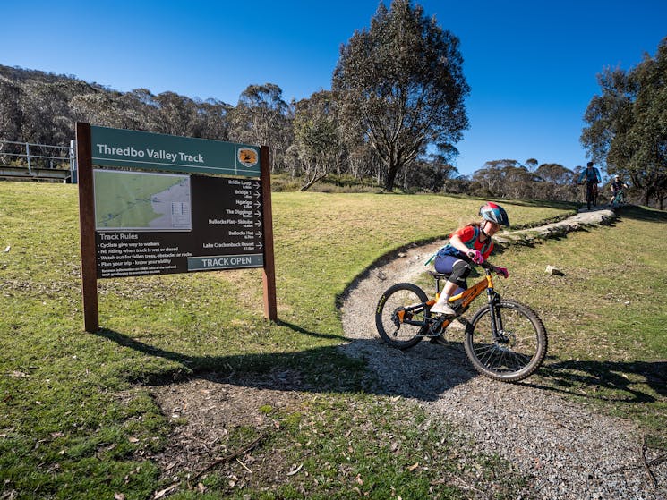 A young girl cycles past Thredbo Valley track signage at the start of the track, in Thredbo