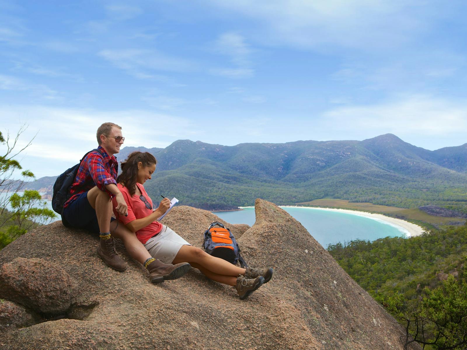 Young hiking couple sitting on huge pink granite boulder on the Hazards,  Wineglass Bay in B/G