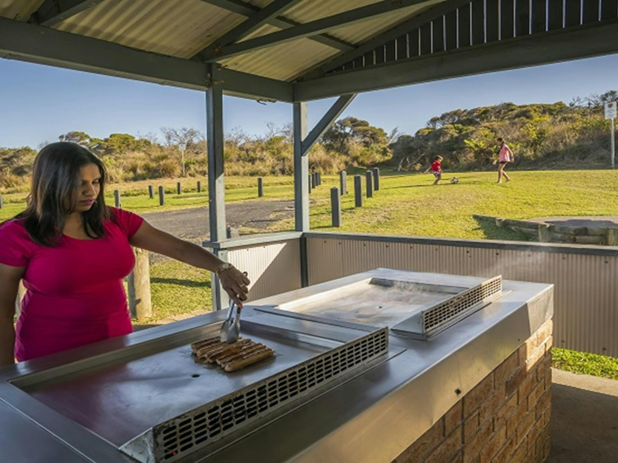 Ein Erwachsener bereitet das Mittagessen auf dem Grill zu, Kinder spielen in der Nähe Fußball, Campingplatz Frazer.