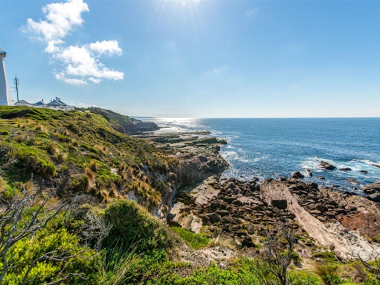 Distant view of Green Cape Lighthouse and outbuildings set on rugged coastal terrain with rocky