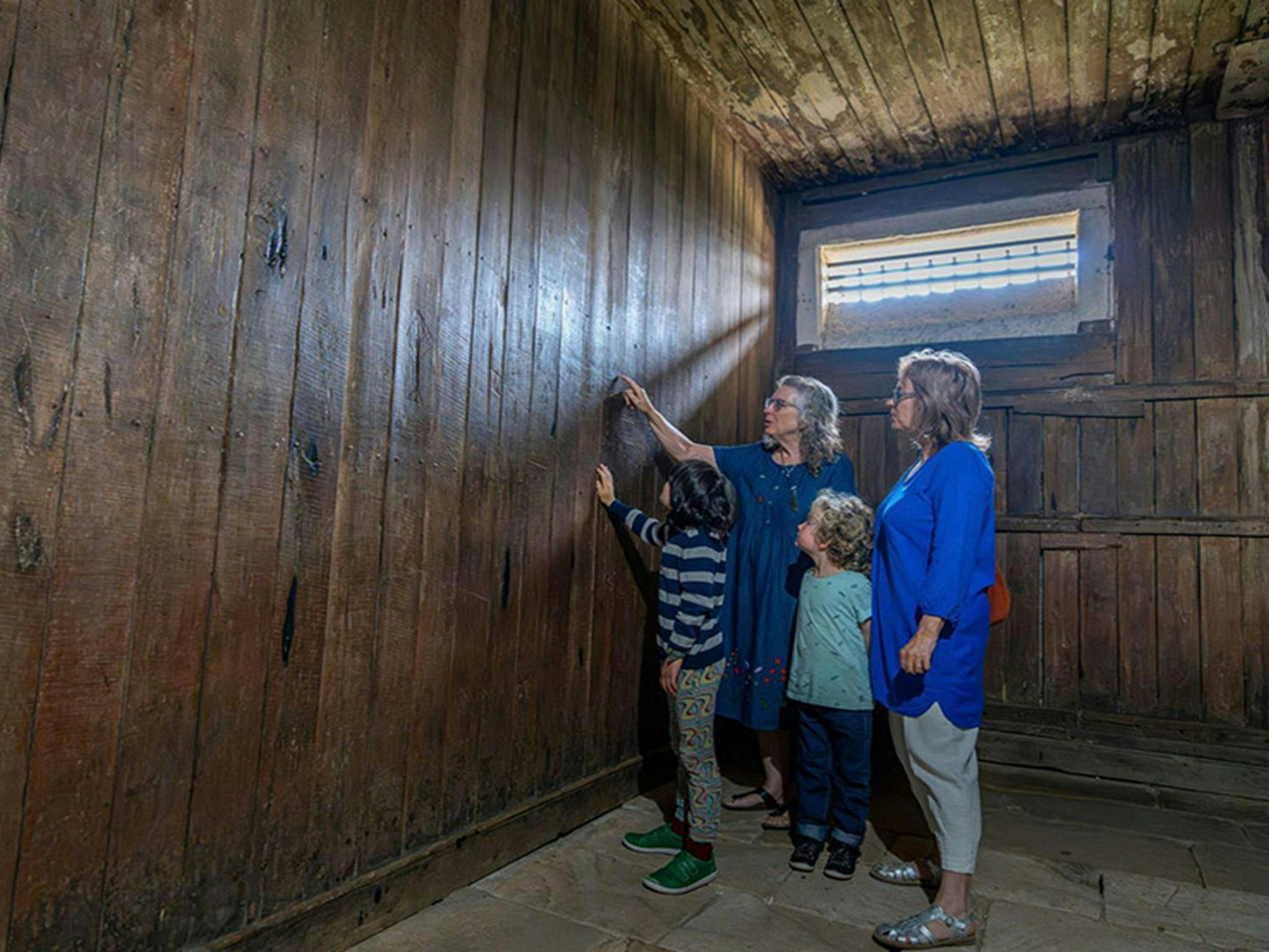 4 visitors inside an old jail cell looking at graffiti left by convicts on the cell walls inside