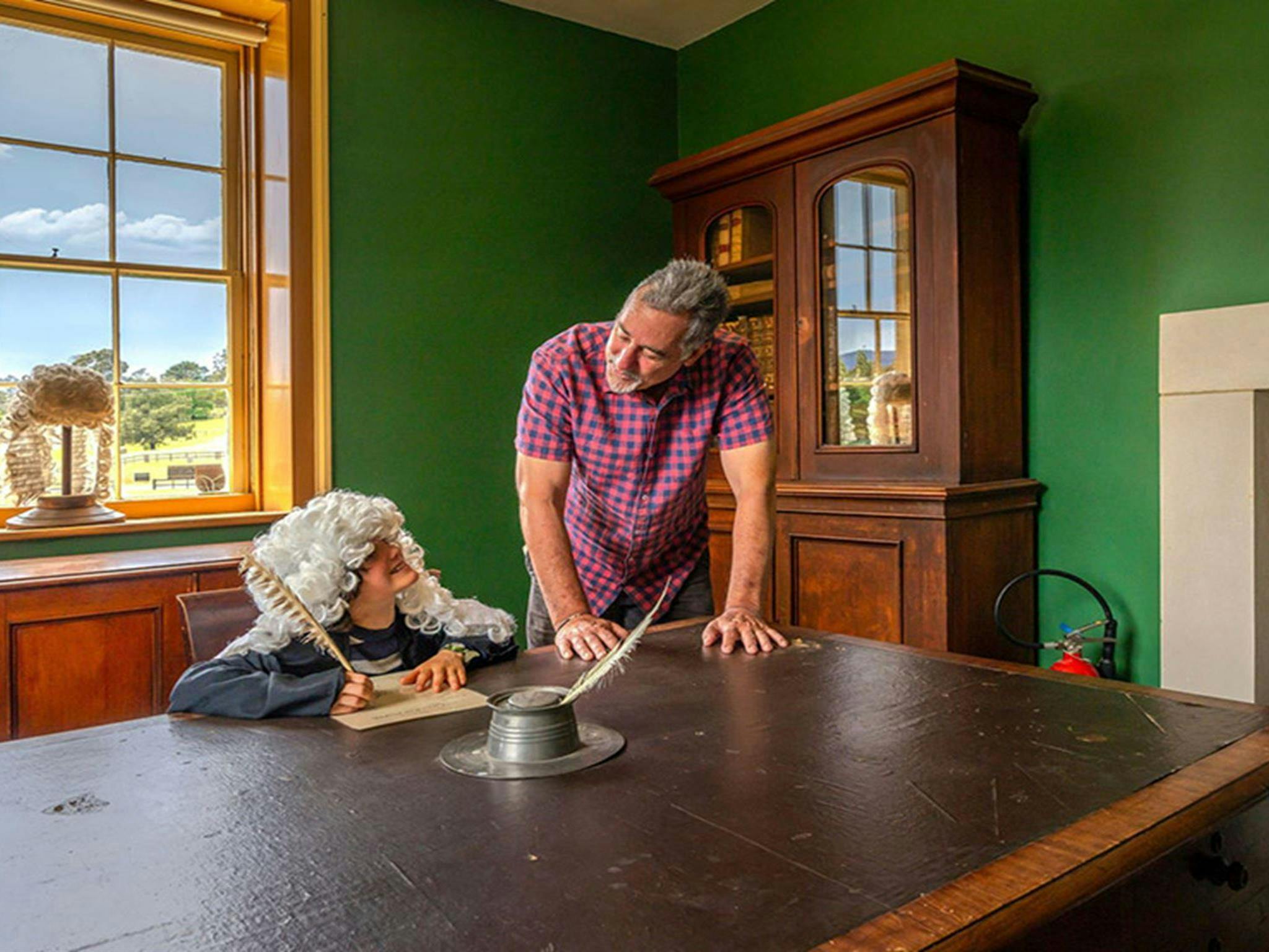 A child dressed as a judge pretending to write with a feather quill with an adult at a desk inside