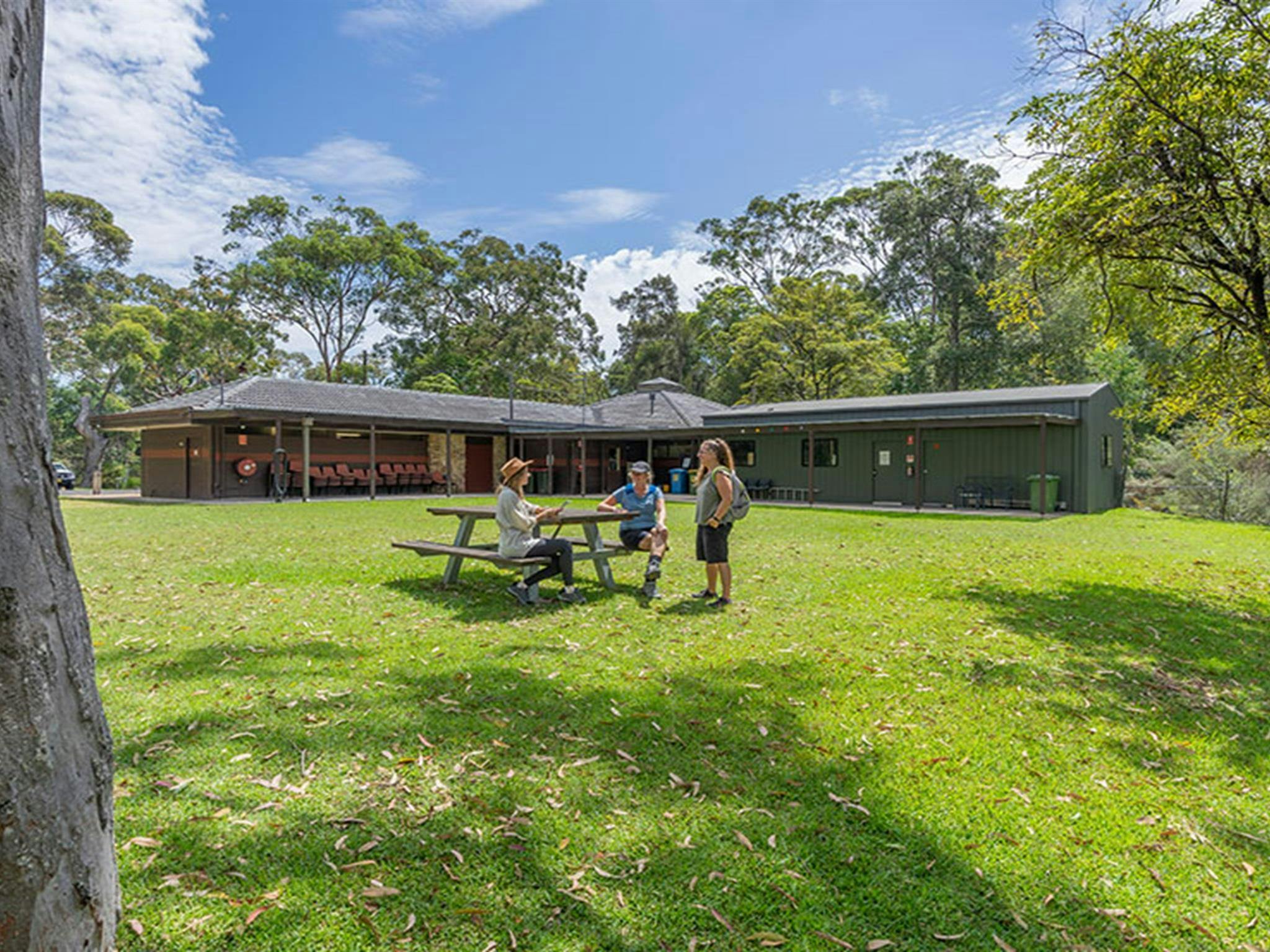 Two people sit on a picnic table bench and one person stands on grassy lawn behind Kalkari Discovery