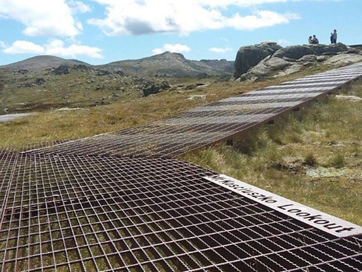 A metal mesh path heading up towards Kosciuszko lookout with mountains in the background in