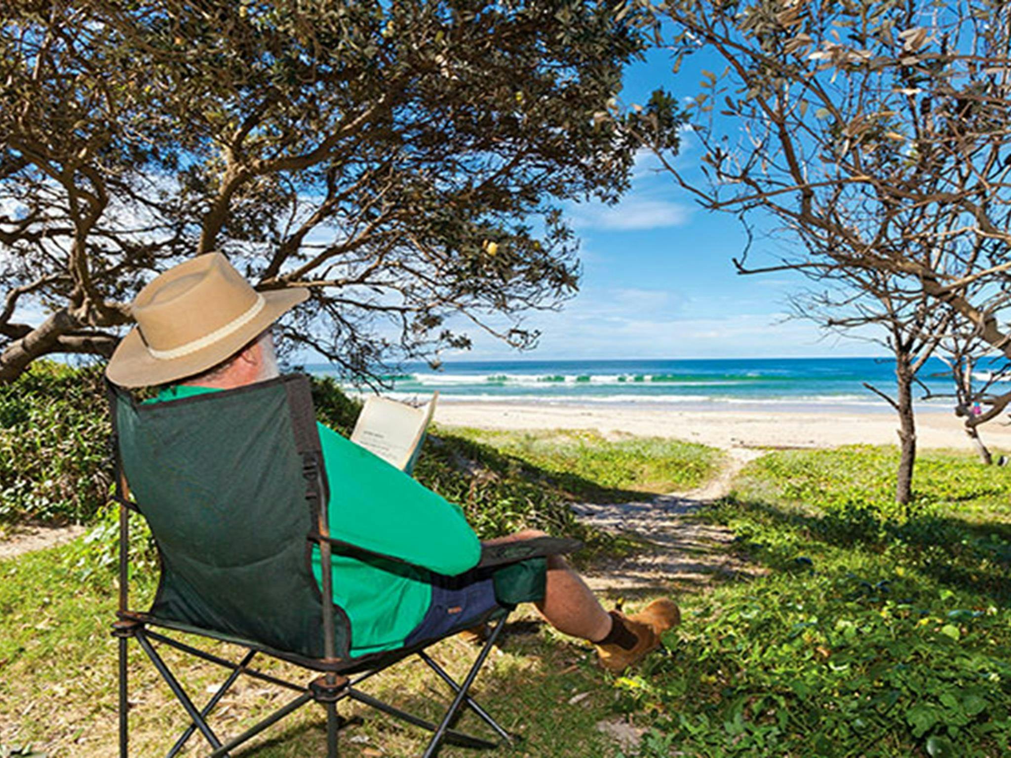 Ein Mann liest im Schatten mit Blick auf den Strand auf dem Campingplatz Red Cliff im Yuraygir-Nationalpark ein Buch.