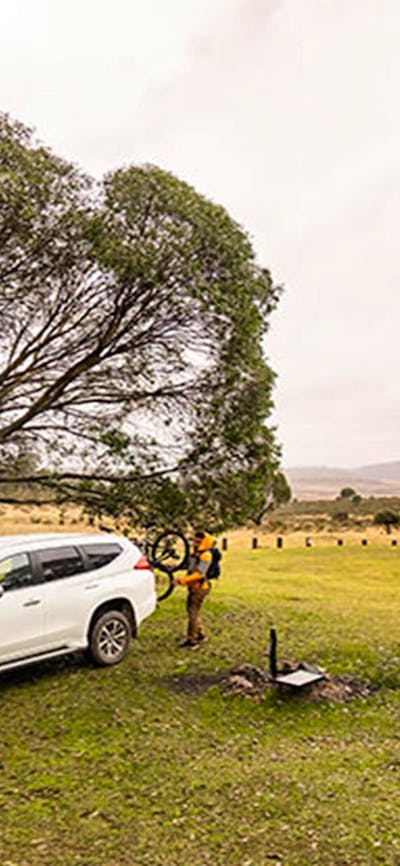 A man sets up a tent next to a fire ring and car at Long Plain Hut campground, Kosciuszko National