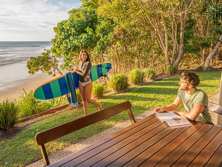 Woman holding a surfboard by the ocean in the backyard of Mildenhall cottage. Photo: DPIE/John