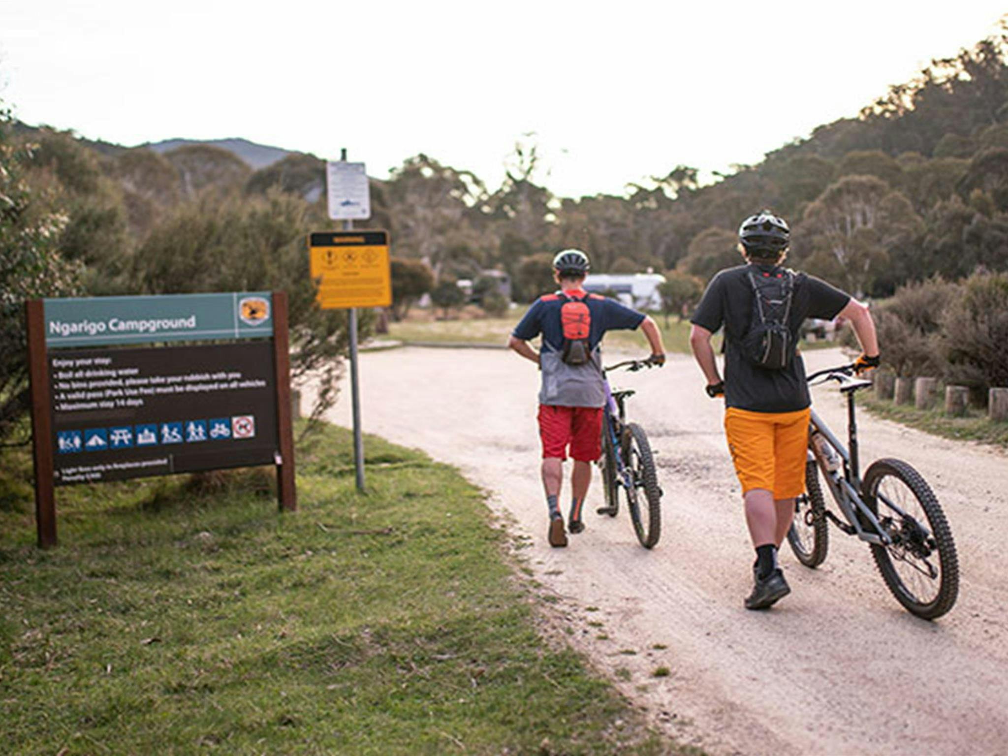 Two mountain bike riders walk past a sign entering Ngarigo campground, Kosciuszko National Park.