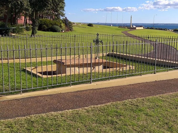Fenced area with tomb and metal cross, on a wide grassy slope with Botany Bay in the background,