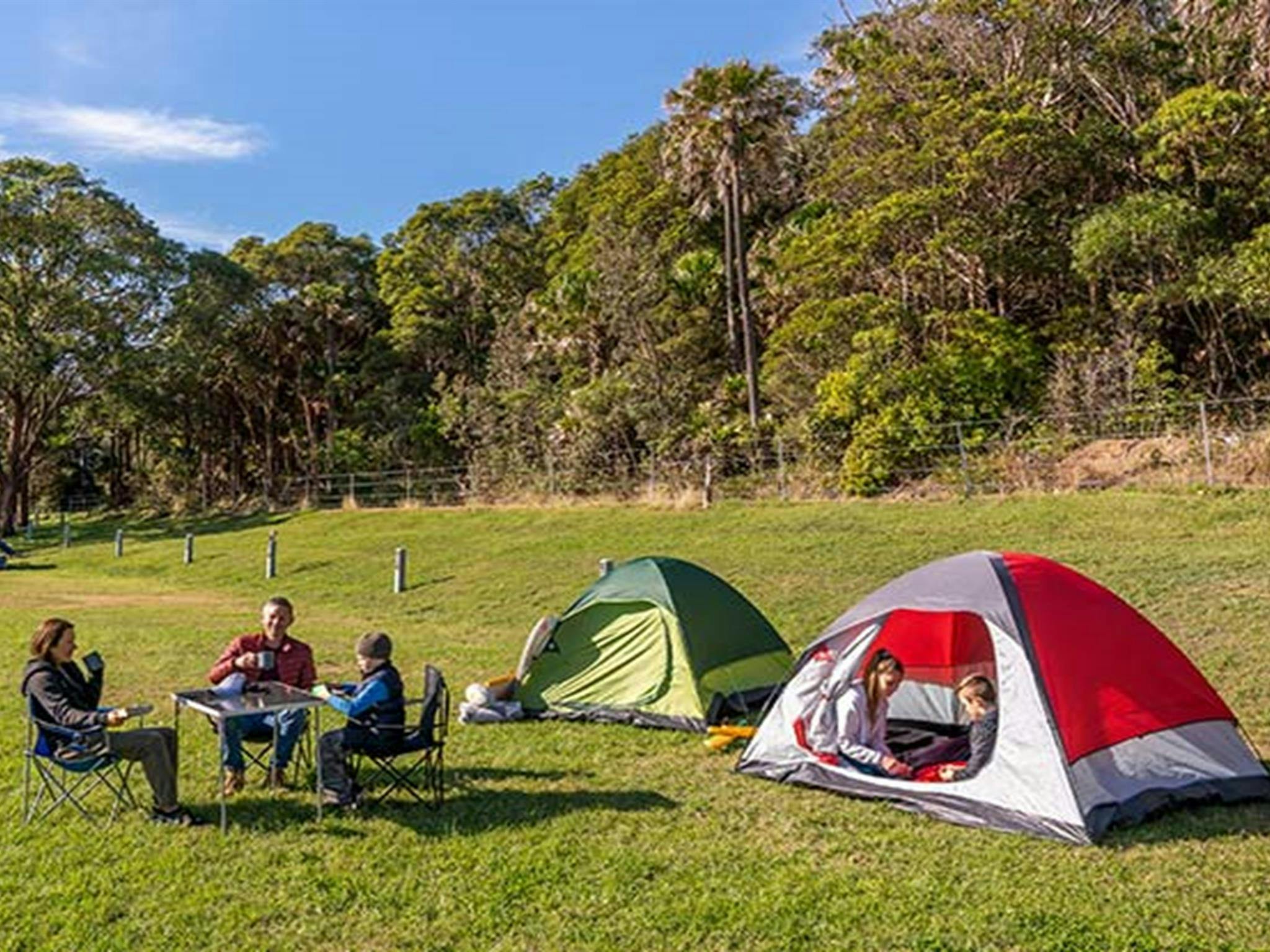 Menschen sitzen an einem Campingtisch und trinken Tee, während ihre Kinder im Zelt spielen. Die Ruinen