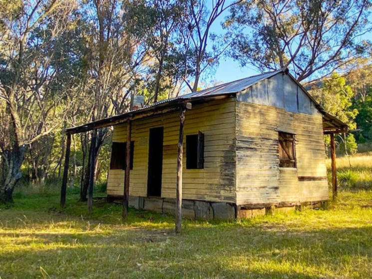 Side view of Scutts Hut set in grassy clearing with hills and bush land in the background. Photo