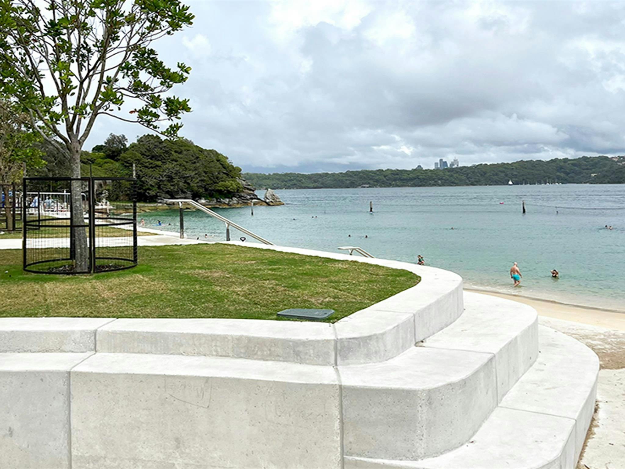 Curved concrete seawall with steps for seating with a grassy patch with trees at the top, and beach