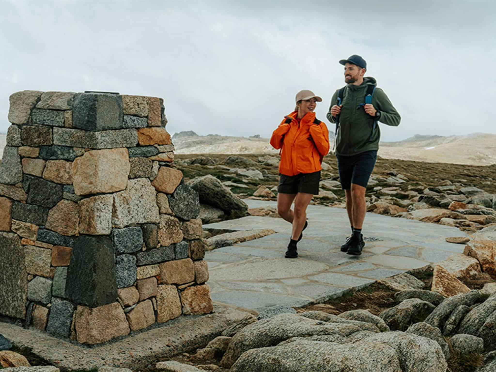 A man and woman walking towards the stone trig at the summit of Mount Kosciuszko with mountain peaks