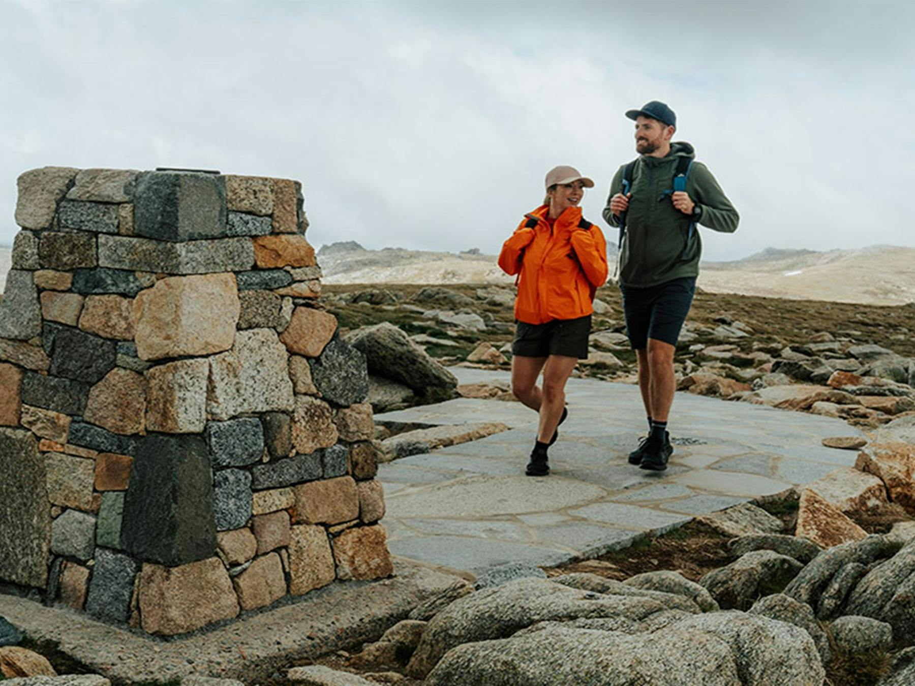 A man and woman walking towards the stone trig at the summit of Mount Kosciuszko with mountain peaks