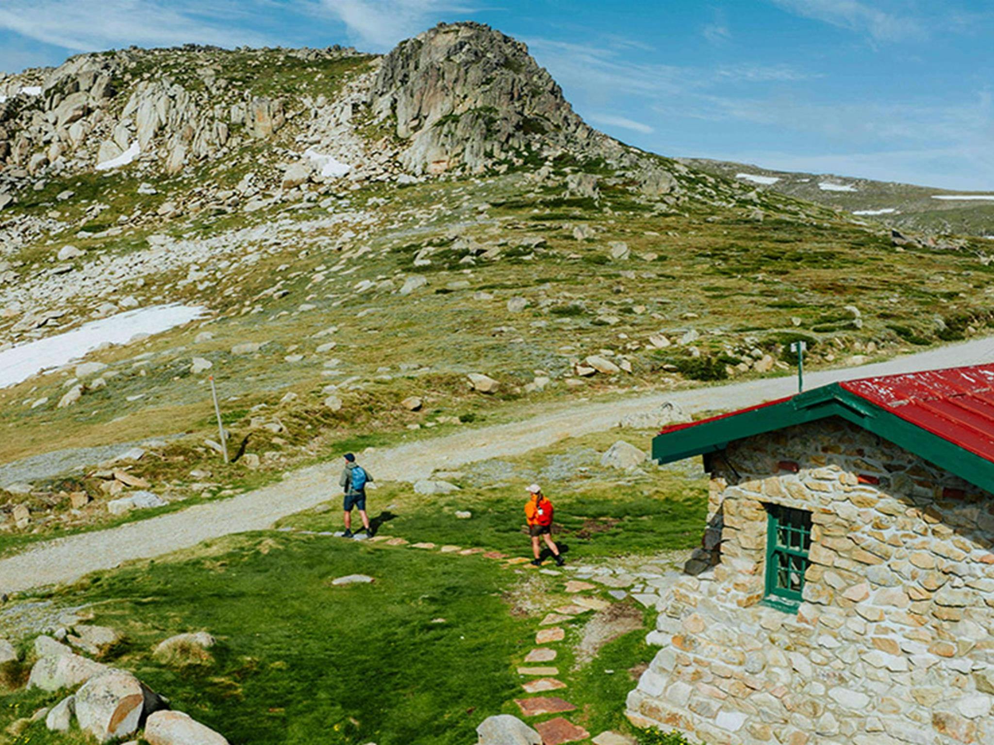 Seamans Hut in the foreground with 2 hikers walking towards the Snowy Mountain peaks of Kosciuszko