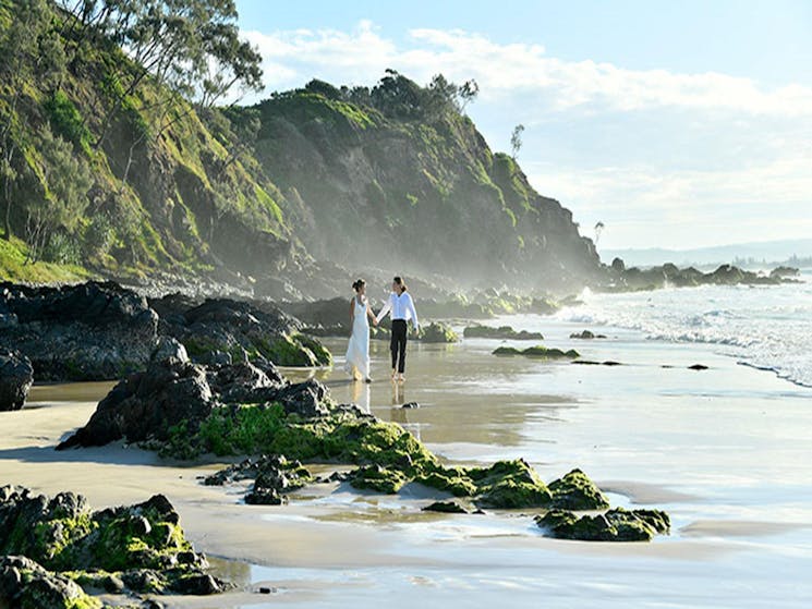 A couple walking past rocks and tidal pools on Wategos Beach in Cape Byron State Conservation Area.
