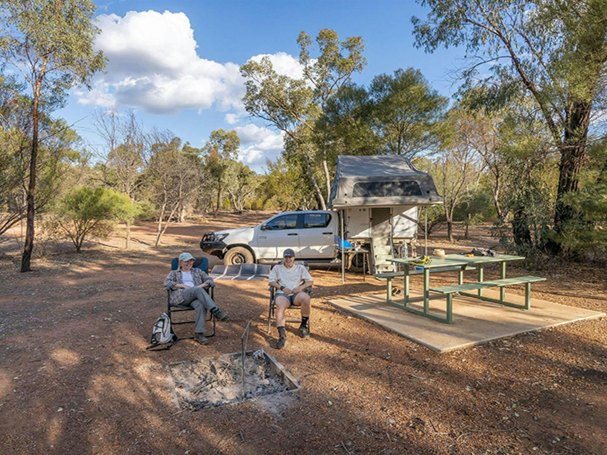 Two people sitting near picnic tables, a campfire and their tent and car at Woolshed Flat campground